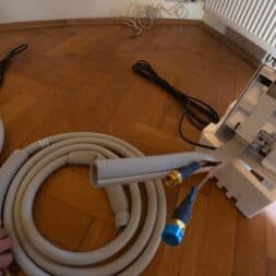 Installing Air Conditioning Yourself in an Older Building – Heating and Cooling in Practice 23 A hand holding an insulated hose near a white air conditioner on a wooden floor, with cables and installation materials scattered around it. www.commaik.de