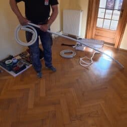 Installing Air Conditioning Yourself in an Older Building – Heating and Cooling in Practice 24 A person is standing in a room with herringbone parquet flooring, holding a coiled cable and a long white hose in their hand—tools and materials are scattered on the floor, suggesting that an air conditioning installation is currently underway. In the background, you can see a radiator, a door, and a window. www.commaik.de
