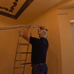 Installing Air Conditioning Yourself in an Older Building – Heating and Cooling in Practice 38 Two people are installing an air conditioner in a room with yellow walls; one is standing on a ladder and connecting pipes, while the other is holding the indoor unit in place from below. Daylight streams in through a window on the right side of the picture. www.commaik.de