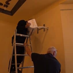Installing Air Conditioning Yourself in an Older Building – Heating and Cooling in Practice 39 Two men are mounting a projector on a beige wall near an air conditioner. One man is standing on a ladder, holding the projector steady, while the other supports the ladder from below. Sunlight streams in through a window on the right. www.commaik.de
