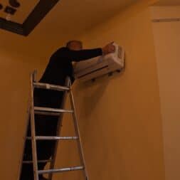 Installing Air Conditioning Yourself in an Older Building – Heating and Cooling in Practice 40 A person standing on a ladder is installing or repairing a wall-mounted air conditioner in a room with yellow walls and a window on the right side. www.commaik.de