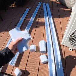 Installing Air Conditioning Yourself in an Older Building – Heating and Cooling in Practice 43 A person is holding a white plastic cover for an air conditioner over several similar parts and long white ducts laid out on a brown wooden deck next to an outdoor air conditioner unit. www.commaik.de
