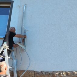 Installing Air Conditioning Yourself in an Older Building – Heating and Cooling in Practice 45 A man standing on a ladder is installing or repairing electrical wiring for an air conditioning unit on the exterior wall of a building, while another person watches from below. www.commaik.de