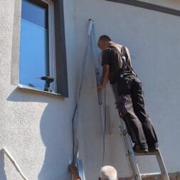 Installing Air Conditioning Yourself in an Older Building – Heating and Cooling in Practice 44 A man is standing on a ladder, installing or repairing a vertical exterior blind or an awning near an air conditioner on the side of the building, while another person assists him from below. A drill is sitting on the windowsill nearby. www.commaik.de