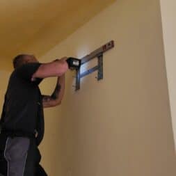 Installing Air Conditioning Yourself in an Older Building – Heating and Cooling in Practice 32 A person is standing on a ladder and using a drill to attach a bracket for an air conditioner to a beige wall. A level rests on the mount to ensure that it is positioned straight, while daylight streams in through a nearby window. www.commaik.de