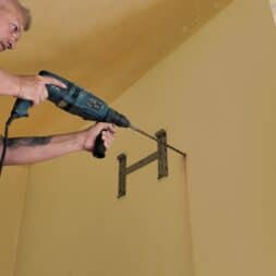 Installing Air Conditioning Yourself in an Older Building – Heating and Cooling in Practice 33 A person standing on a ladder is using a drill to attach metal brackets to a yellow wall in preparation for installing an air conditioner. www.commaik.de