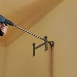 Installing Air Conditioning Yourself in an Older Building – Heating and Cooling in Practice 34 A person standing on a ladder is using a drill and a long drill bit to drill a hole in a beige wall to install an air conditioner, leaving a vertical trail of dust below a metal bracket on the wall. www.commaik.de