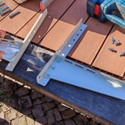 Installing Air Conditioning Yourself in an Older Building – Heating and Cooling in Practice 42 A person is using a wrench to attach metal brackets—possibly for the installation of an air conditioner—to a wooden surface outdoors, with screws and tools lying nearby. www.commaik.de