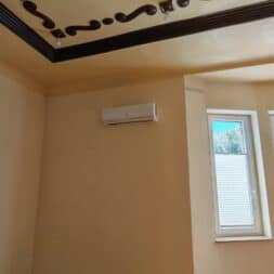 Installing Air Conditioning Yourself in an Older Building – Heating and Cooling in Practice 59 A beige room with two windows, partially covered by white blinds, a wall-mounted air conditioner, and a decorative ceiling molding featuring wavy dark patterns and small lights. www.commaik.de
