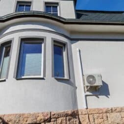 Installing Air Conditioning Yourself in an Older Building – Heating and Cooling in Practice 48 A modern house facade with three rectangular windows, beige walls, a gray tile roof, and an air conditioner mounted on the exterior wall above a stone base. The sky is clear and blue. www.commaik.de