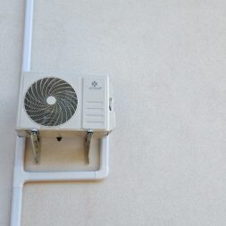Installing Air Conditioning Yourself in an Older Building – Heating and Cooling in Practice 49 A wall-mounted outdoor air conditioning unit with a visible fan and pipes is installed on a light-colored exterior wall near a window. www.commaik.de