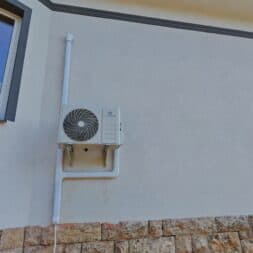 Installing Air Conditioning Yourself in an Older Building – Heating and Cooling in Practice 6 A wall-mounted air conditioner is installed on the exterior wall of a house that has a light-colored wall, a large window, and a stone section at the bottom. www.commaik.de