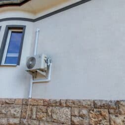 Installing Air Conditioning Yourself in an Older Building – Heating and Cooling in Practice 50 An air conditioner is mounted on an exterior wall of a building with a stone base and a white upper structure, next to a window and beneath a curved roof. The blue sky can be seen in the background. www.commaik.de
