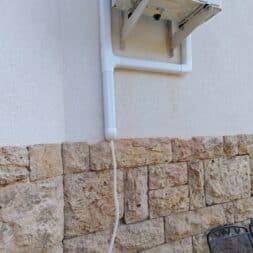 Installing Air Conditioning Yourself in an Older Building – Heating and Cooling in Practice 51 High up on a wall made of plaster and stone, there is an air conditioner with a white drain pipe running down from it. On the floor next to the wall are a metal chair and a few logs. www.commaik.de