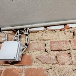 Installing Air Conditioning Yourself in an Older Building – Heating and Cooling in Practice 57 A white junction box for the air conditioning system, with labeled cables, is mounted on a rough, unplastered brick wall directly below a white ceiling. Several white cables run along the wall and ceiling. www.commaik.de