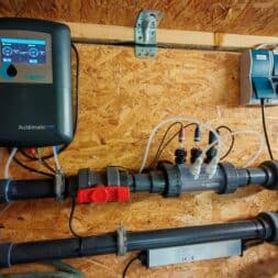 Installing the BAYROL Automatic Cl-pH in the Pool and Retrofitting a Temperature Sensor: My Experience 5 Attached to a wooden wall are an automatic Bayrol water monitoring and control panel and valves connected to each other by black pipes, with hoses and electronic components visible. www.commaik.de