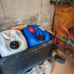 Installing the BAYROL Automatic Cl-pH in the Pool and Retrofitting a Temperature Sensor: My Experience 14 Two plastic containers, one white and one blue, are sitting in a black box against a wall, near which there are pipes, cables, and valves—in a utility or maintenance room, such as those often found near automatic Bayrol pool care systems. www.commaik.de