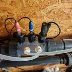 High Chlorine Consumption in the Pool: Why 20 Liters of Chlorine Disappeared in Two Weeks 5 Close-up of a plumbing system with hoses, connectors, and sensors attached to gray pipes, which are in turn mounted on a plywood wall. The system includes colored cables and labels on the components for monitoring flow rates and chlorine consumption. www.commaik.de