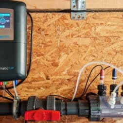Installing the BAYROL Automatic Cl-pH in the Pool and Retrofitting a Temperature Sensor: My Experience 12 Bayrol automatic pool dosing unit with a labeled display and a transparent housing, connected via hoses and cables; mounted on a wooden wall, with gray pipes and a red valve visible below it. www.commaik.de