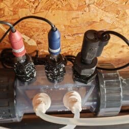 Installing the BAYROL Automatic Cl-pH in the Pool and Retrofitting a Temperature Sensor: My Experience 17 Three electronic sensors with black cables are attached to a gray PVC pipe secured to a wooden wall; two white hoses are connected at the bottom—an arrangement commonly found in automatic Bayrol systems for precise water quality control. www.commaik.de