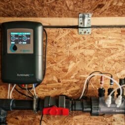 High Chlorine Consumption in the Pool: Why 20 Liters of Chlorine Disappeared in Two Weeks 4 Mounted on a wooden wall above the plumbing pipes and valves is a black automatic control unit with a digital display that monitors water flow and chlorine consumption in a filtration or irrigation system. www.commaik.de