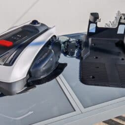 ECOVACS GOAT O1200 LiDAR PRO Review: Perfect Edge Mowing Without Any Touch-Ups? 7 An Ecovacs Goat robotic lawn mower, a charging station, and a power adapter are sitting on a glass outdoor table in the sunlight. In the background, you can see a potted plant and a wire fence. www.commaik.de