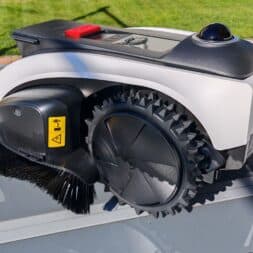 ECOVACS GOAT O1200 LiDAR PRO Review: Perfect Edge Mowing Without Any Touch-Ups? 9 An Ecovacs Goat robotic lawn mower with large wheels and a side brush is sitting on a glass table outdoors, near a lawn and a paved area. You can also see an instruction manual and a cable on the table. www.commaik.de