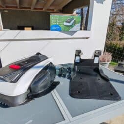 ECOVACS GOAT O1200 LiDAR PRO Review: Perfect Edge Mowing Without Any Touch-Ups? 10 A robotic lawn mower, its charging station, and accessories are on display on an outdoor table. On a counter nearby is a package of the Ecovacs Goat, and a modern garden setting can be seen in the background. www.commaik.de
