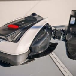 ECOVACS GOAT O1200 LiDAR PRO Review: Perfect Edge Mowing Without Any Touch-Ups? 11 An Ecovacs Goat robotic lawn mower with brushes is sitting on a table next to its charging station and power adapter, outdoors in bright sunlight. www.commaik.de