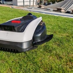 ECOVACS GOAT O1200 LiDAR PRO Review: Perfect Edge Mowing Without Any Touch-Ups? 3 An ECOVACS Goat robotic lawn mower with a black-and-white body is driving across a green lawn on a sunny day; a building and some covered structures can be seen in the background. www.commaik.de