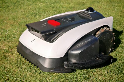 ECOVACS GOAT O1200 LiDAR PRO Review: Perfect Edge Mowing Without Any Touch-Ups? 4 An Ecovacs Goat robotic lawn mower with a black-and-white body stands in the sunlight on a green lawn; it has a red button and a front brush. www.commaik.de