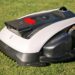 ECOVACS GOAT O1200 LiDAR PRO im Test: Perfektes Kantenmähen ohne Nacharbeit? 59 Ein Ecovacs Goat-Mähroboter mit weiß-schwarzem Gehäuse steht im Sonnenlicht auf grünem Rasen; er verfügt über einen roten Knopf und eine Frontbürste. www.commaik.de