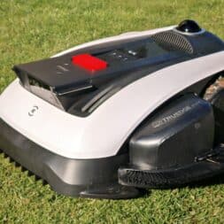 ECOVACS GOAT O1200 LiDAR PRO Review: Perfect Edge Mowing Without Any Touch-Ups? 4 A robotic lawn mower with a black-and-white body stands on a green lawn in daylight; a red button and a visible brush can be seen on its side—the design is reminiscent of the elegant Ecovacs Goat series. www.commaik.de