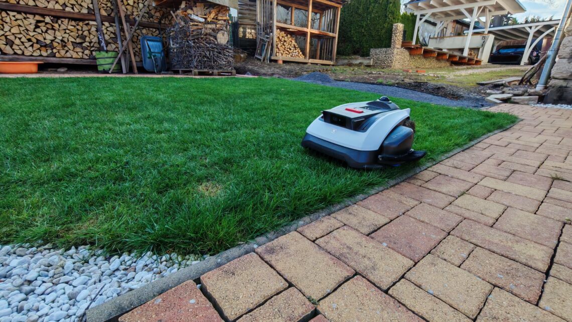 Garten 1 Ein Mähroboter ist auf einem grünen Rasen in der Nähe eines Steinwegs im Einsatz, im Hintergrund sind gestapeltes Brennholz und Holzkonstruktionen zu sehen. Dieser Mähroboter nutzt fortschrittliche LiDAR-Technologie zur präzisen Navigation, sodass kein Begrenzungskabel erforderlich ist. www.commaik.de