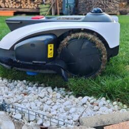ECOVACS GOAT O1200 LiDAR PRO Review: Perfect Edge Mowing Without Any Touch-Ups? 5 An Ecovacs Goat robotic lawn mower with grass-covered wheels is standing at the edge of a green lawn and a garden border planted with stones. In the background, you can see stacks of firewood and gardening tools. www.commaik.de