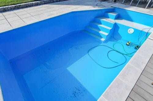 High Chlorine Consumption in the Pool: Why 20 Liters of Chlorine Disappeared in Two Weeks 20 An almost empty swimming pool with some dirt on the bottom, a green hose, and a small pump. A staircase leads down to the pool, which is surrounded by light-colored tiles—factors that can affect chlorine usage when you’re preparing the pool for use. www.commaik.de