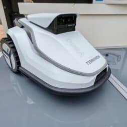 TerraMow V1000, V600, and Mini 500 Review: Robotic Lawn Mowers Without RTK and Lidar? 15 A white, modern TERRAMON robotic lawn mower, the Terramow V600, is parked outdoors on a reflective surface near a garden and patio area, showcasing the sleek design also found in the advanced Terramow V1000 model. www.commaik.de