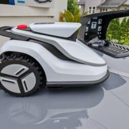 TerraMow V1000, V600, and Mini 500 Review: Robotic Lawn Mowers Without RTK and Lidar? 14 A Terramow V1000 robotic lawn mower with large wheels is standing outdoors on a reflective surface near a house and some green spaces; behind it, its charging station is open. www.commaik.de