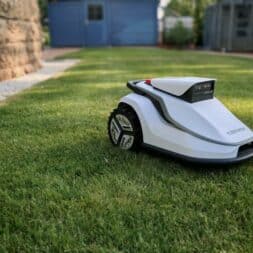 TerraMow V1000, V600, and Mini 500 Review: Robotic Lawn Mowers Without RTK and Lidar? 11 A white Terramow V1000 robotic lawn mower moves effortlessly across a well-manicured green lawn in a backyard; in the background, a blue shed and lush plants are visible. www.commaik.de