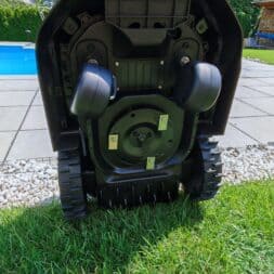 TerraMow V1000, V600, and Mini 500 Review: Robotic Lawn Mowers Without RTK and Lidar? 31 The underside of a Terramow V1000 robotic lawn mower is visible on a lawn near a pool, with its wheels, blades, and mechanical parts clearly visible in bright daylight. www.commaik.de