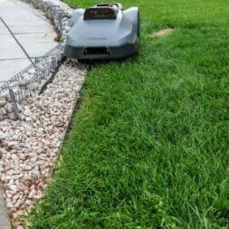 TerraMow V1000, V600, and Mini 500 Review: Robotic Lawn Mowers Without RTK and Lidar? 35 A Terramow V1000 robotic lawn mower is mowing the grass near a stone border in a backyard; in the background, you can see stacked firewood and patio furniture. www.commaik.de