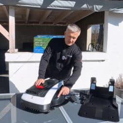 ECOVACS GOAT O1200 LiDAR PRO Review: Perfect Edge Mowing Without Any Touch-Ups? 13 A man is standing outside at a glass table, setting up or checking an Ecovacs Goat robotic lawn mower and its charging station near a covered patio. www.commaik.de