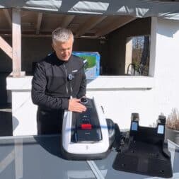 ECOVACS GOAT O1200 LiDAR PRO Review: Perfect Edge Mowing Without Any Touch-Ups? 15 A man is standing outdoors next to a glass table, operating an Ecovacs Goat robot, next to which is a black charging station. A covered patio and a garden can be seen in the background. www.commaik.de