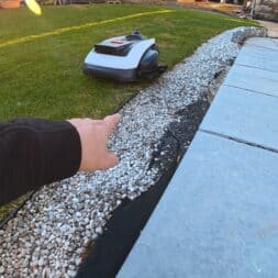 ECOVACS GOAT O1200 LiDAR PRO Review: Perfect Edge Mowing Without Any Touch-Ups? 46 A person's outstretched arm points toward an Ecovacs Goat robotic lawn mower working on a green lawn next to a strip of white stones and a paved area in a backyard. www.commaik.de
