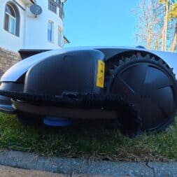 ECOVACS GOAT O1200 LiDAR PRO Review: Perfect Edge Mowing Without Any Touch-Ups? 53 A close-up of an Ecovacs Goat robotic lawn mower on a lawn near a house, with trees and a bright blue sky visible in the background. www.commaik.de