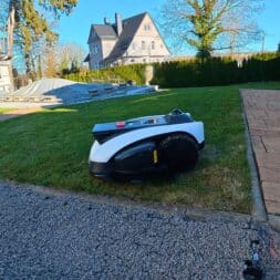 ECOVACS GOAT O1200 LiDAR PRO Review: Perfect Edge Mowing Without Any Touch-Ups? 45 An Ecovacs Goat robotic lawn mower is driving across green grass near a paved walkway in a sunny garden; a house, trees, and a pile of wood can be seen in the background. www.commaik.de