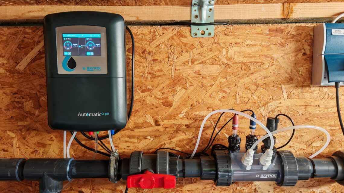 Pool 3 A digital BAYROL Automatic Cl-pH controller for swimming pools and the water treatment system connected to it are mounted on a wooden wall, with pipes, valves, and transparent sections for monitoring water flow and chemical dosing are visible, ensuring precise installation and efficient chemical management. www.commaik.de