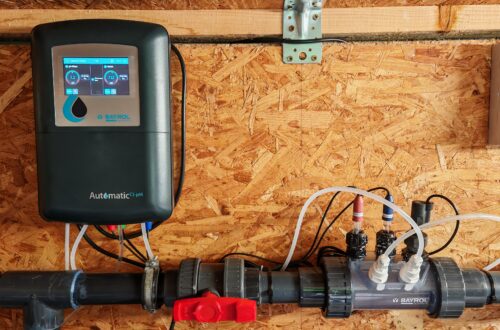 BAYROL Automatic Cl-pH: Installation and Field Test A digital BAYROL Automatic Cl-pH controller for swimming pools and the water treatment system connected to it are mounted on a wooden wall, with pipes, valves, and transparent sections for monitoring water flow and chemical dosing are visible, ensuring precise installation and efficient chemical management. www.commaik.de