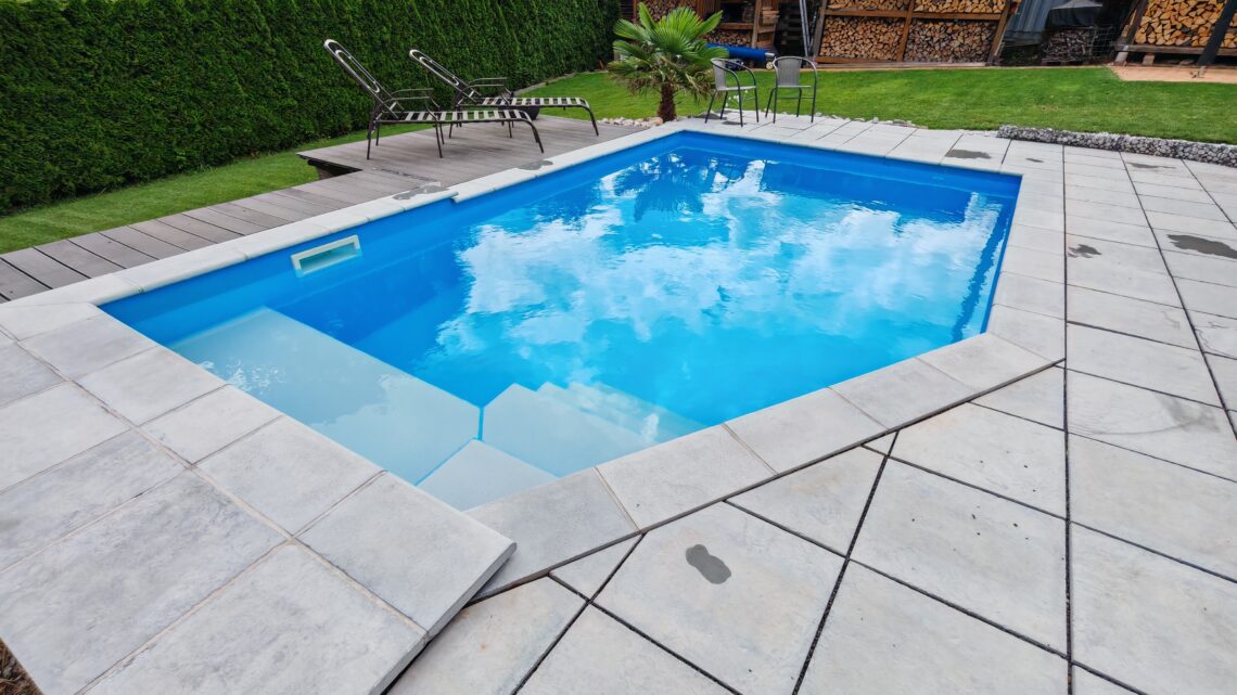 Pool 1 A small, rectangular outdoor pool with clear, blue, chlorinated water is surrounded by light gray tiles. Next to them are two lounge chairs, a small table, and green grass. In the background, you can see shrubs and a pile of firewood. www.commaik.de