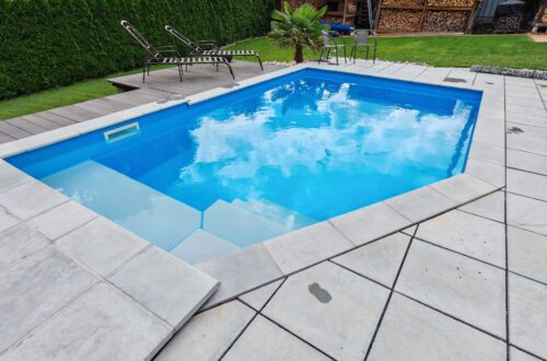 High Chlorine Consumption—20 Liters of Chlorine in Two Weeks—My Troubleshooting at the Pool A small, rectangular outdoor pool with clear, blue, chlorinated water is surrounded by light gray tiles. Next to them are two lounge chairs, a small table, and green grass. In the background, you can see shrubs and a pile of firewood. www.commaik.de